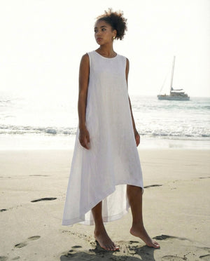 Woman standing on a beach wearing a loose-fitting white linen dress with an asymmetrical hem.