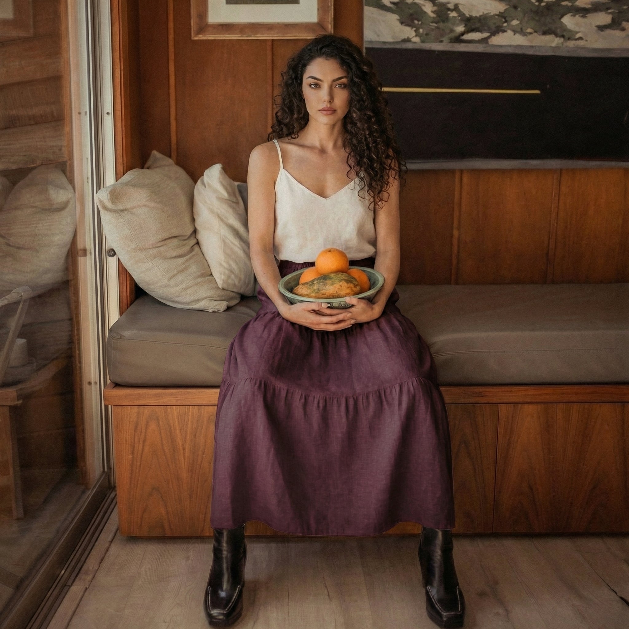 Woman sitting on a wooden bench holding a bowl of oranges, wearing a grape wine linen tiered skirt and black boots.