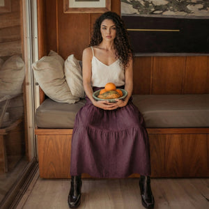 Woman sitting on a wooden bench holding a bowl of oranges, wearing a grape wine linen tiered skirt and black boots.
