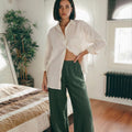 Woman wearing MINA wide leg linen pants in smoke pine green, styled with a relaxed white linen shirt in a bedroom setting.