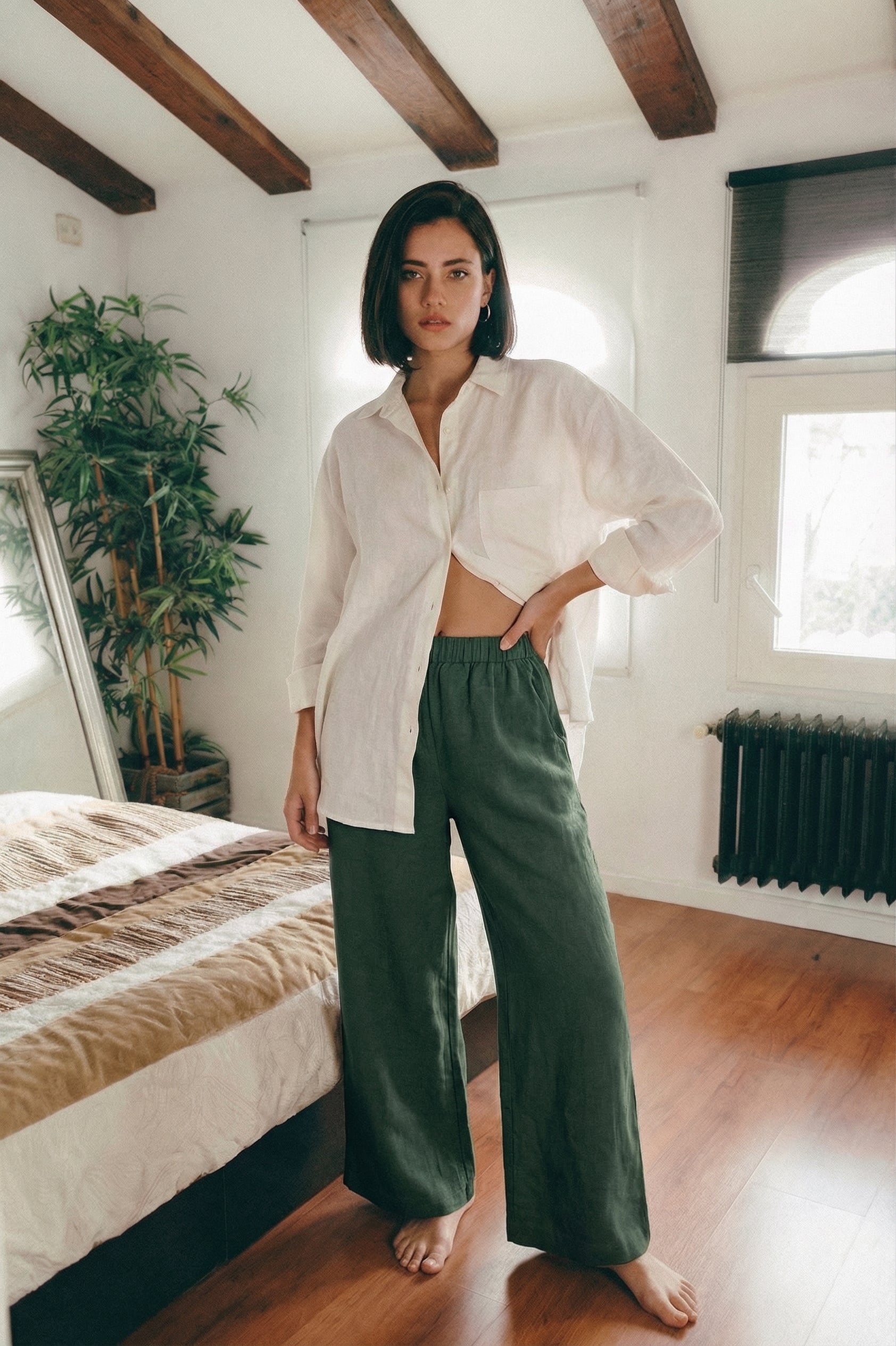 Woman wearing MINA wide leg linen pants in smoke pine green, styled with a relaxed white linen shirt in a bedroom setting.