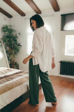 Side and back view of a woman wearing MINA smoke pine wide leg linen pants and an oversized white shirt, standing barefoot in a cozy bedroom.