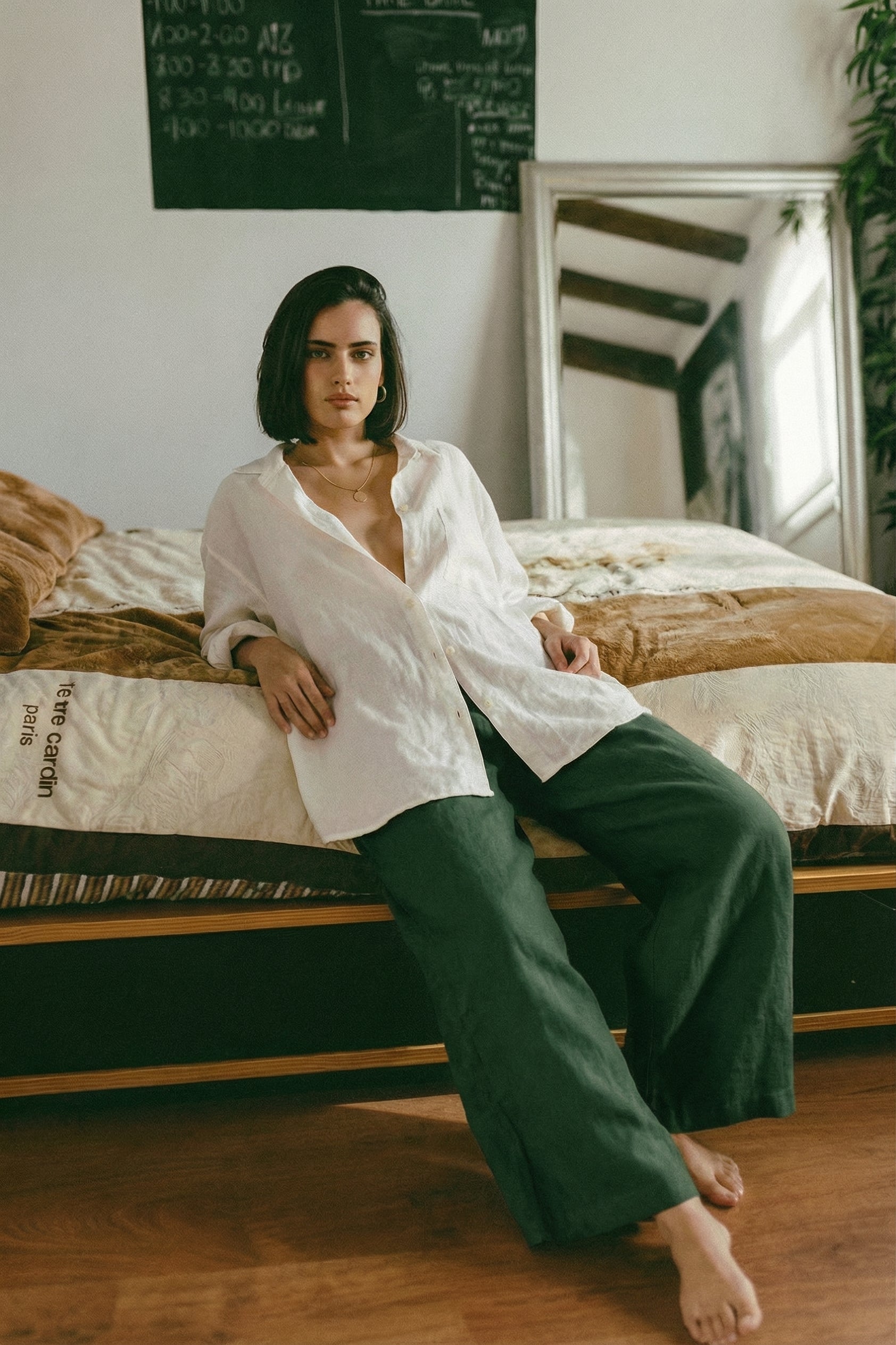 Woman leaning comfortably against a bed, wearing MINA smoke pine wide leg linen pants and a relaxed white shirt, showcasing the flowy fabric.