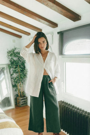 Woman styling MINA smoke pine wide leg linen pants with a breezy, open white shirt, resting one hand in her pocket while standing in a bright room.