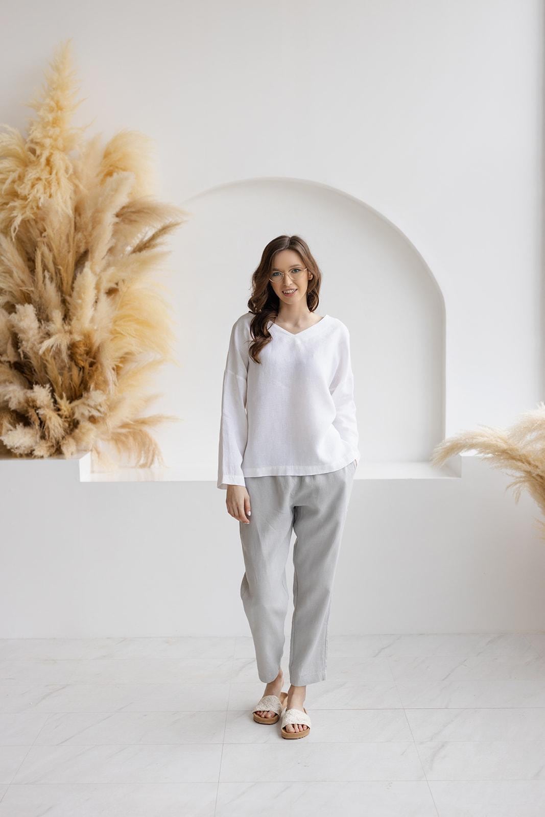 Woman in Solvia white linen blouse and gray linen pants, V-neck, long sleeves, relaxed fit, minimalist studio with pampas grass.