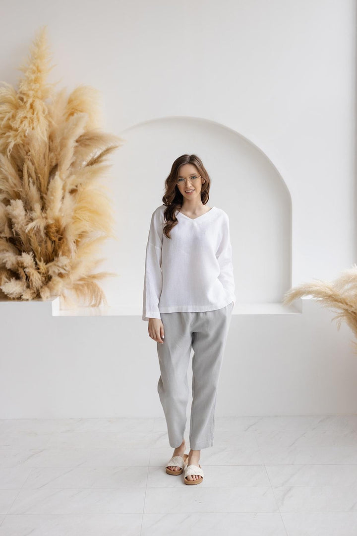 Woman in Solvia white linen blouse and gray linen pants, V-neck, long sleeves, relaxed fit, minimalist studio with pampas grass.