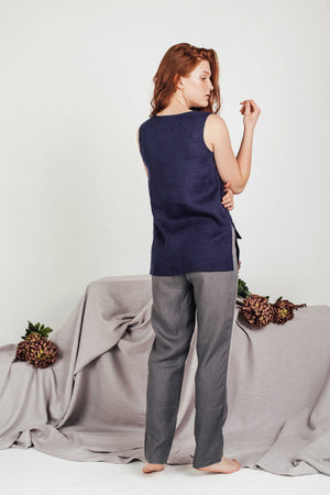 Back view of a model wearing a handmade navy linen top and tailored grey pants, posing barefoot in front of a soft, elegant setting with botanical props.