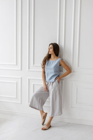 Woman wearing a dusty blue sleeveless linen blouse and light grey wide-leg pants, leaning casually against a white wall in natural light.
