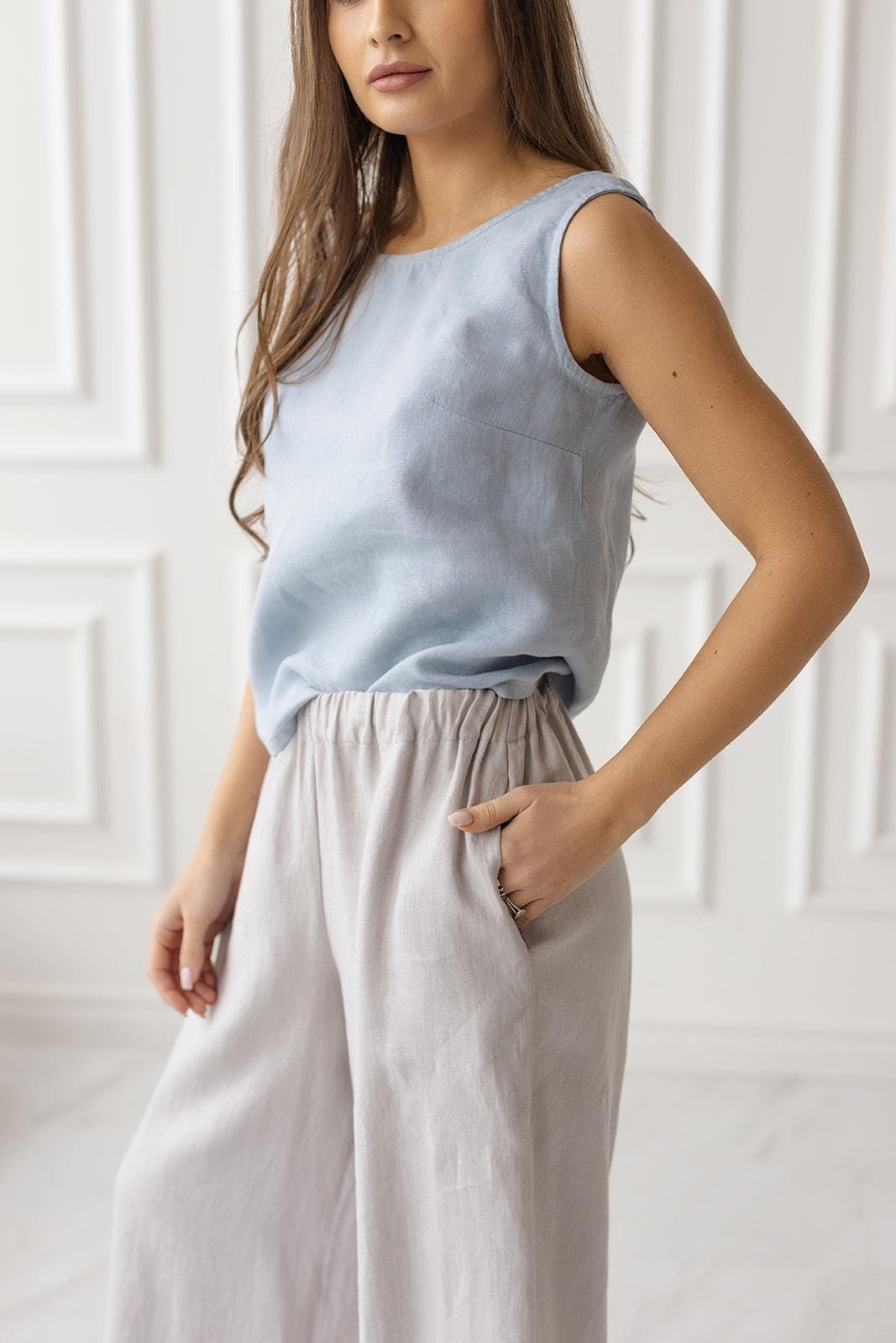 Front view of a model wearing a dusty blue sleeveless linen blouse with a boat neckline, paired with light gray wide-leg linen pants, showcasing a relaxed summer outfit.