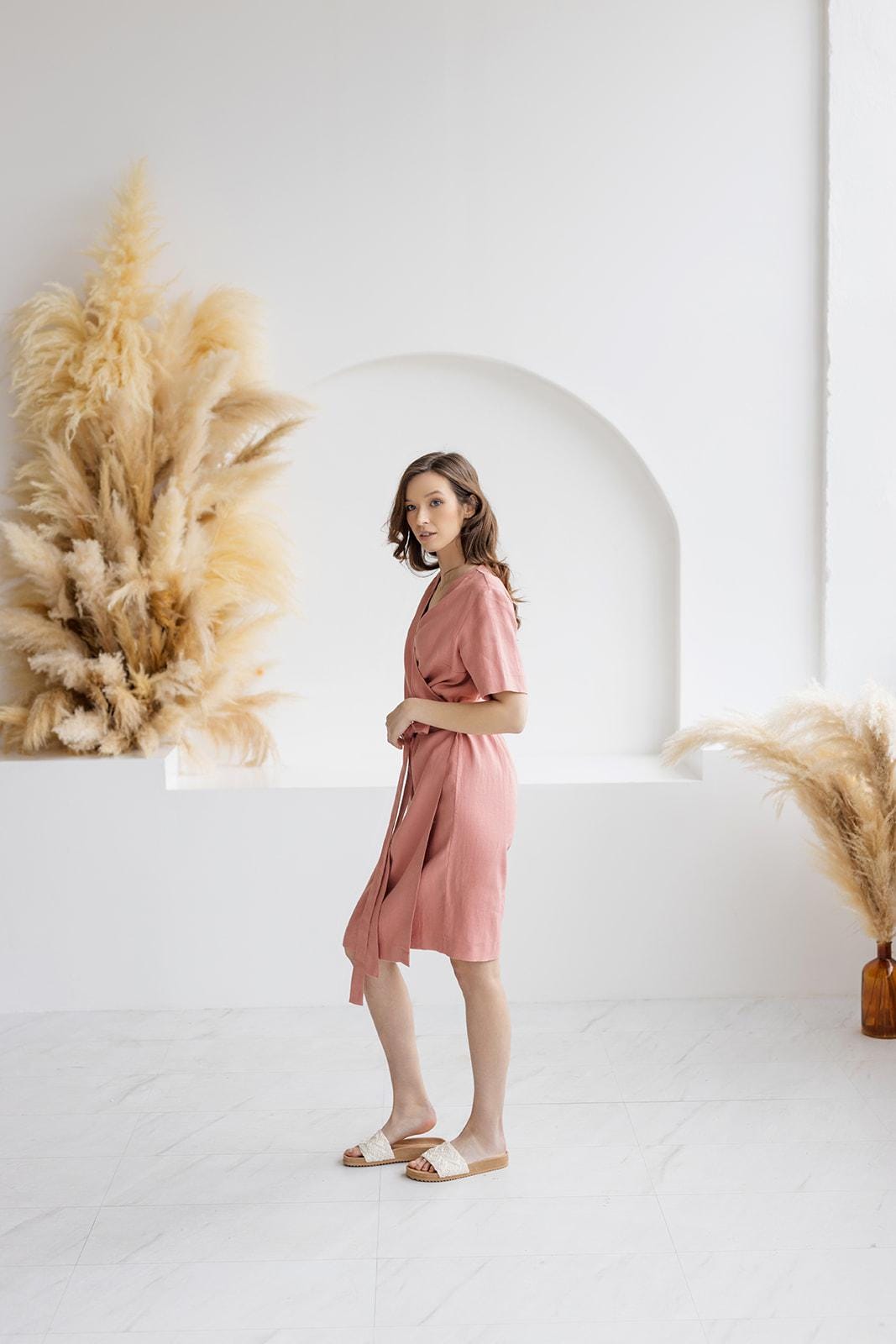 Woman wearing a blush salmon linen wrap dress with a belted waist, V-neckline, and short sleeves. A versatile, breathable, medium-weight linen dress, perfect for casual and summer wear.