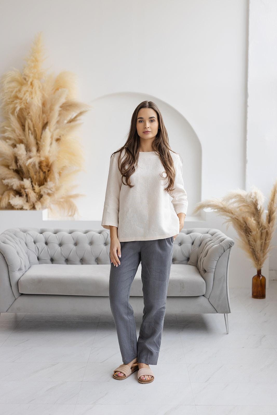 Woman wearing dark gray linen pants and a cream linen top, standing in a modern minimalist living room with a tufted sofa and pampas grass decor.