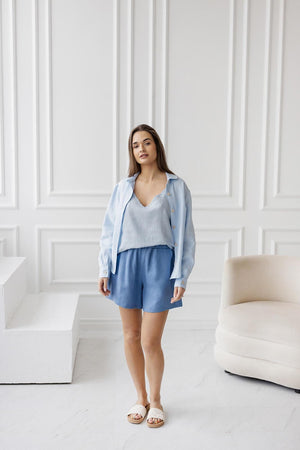 Woman wearing a light blue linen outfit with a V-neck cami, matching shorts, and an open button-down shirt, standing in a minimalist white interior.