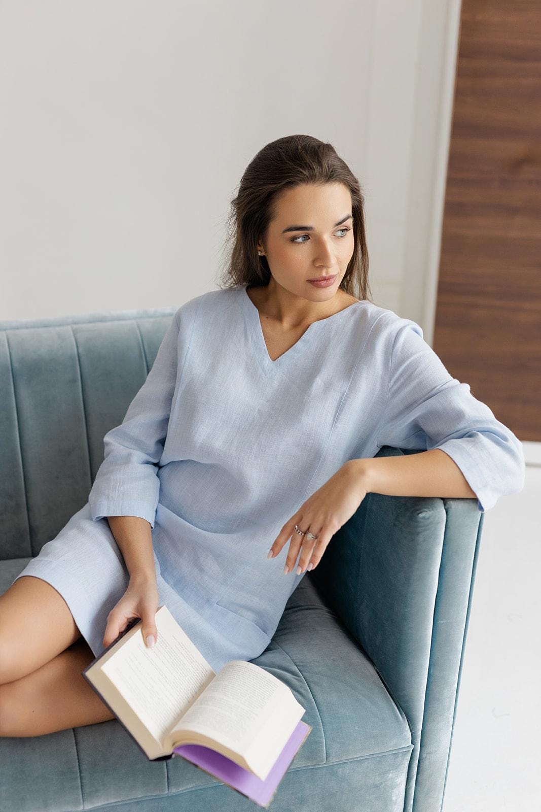 Woman wearing light blue linen shift dress, reading a book on a blue velvet sofa.