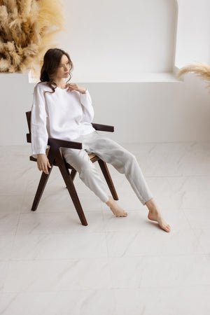 Model seated on wooden chair, wearing dusty gray straight-leg relaxed fit linen pants and white linen top.