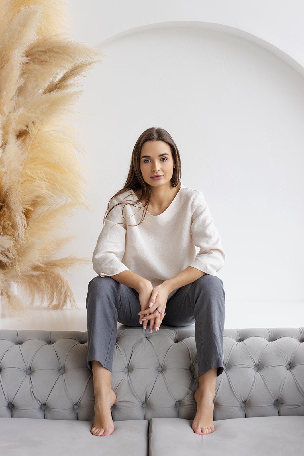 Woman sitting barefoot on a tufted gray sofa, wearing relaxed-fit dark gray linen pants and a soft linen top in a cozy, neutral-toned room.