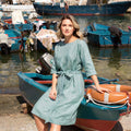 Model wearing a breathable sea-green knee-length linen dress, sitting on a small wooden boat with a sunny Mediterranean harbor in the background.