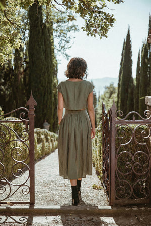 Back view of a model wearing the Sonfre NAOMI eucalyptus green linen crop top and matching high-waisted midi skirt, walking through an ornate iron gate into a garden.