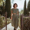 Model wearing the Sonfre NAOMI eucalyptus green linen two-piece set, featuring a boxy crop top and a high-waisted midi skirt, walking through an ornate iron gate in a garden setting.