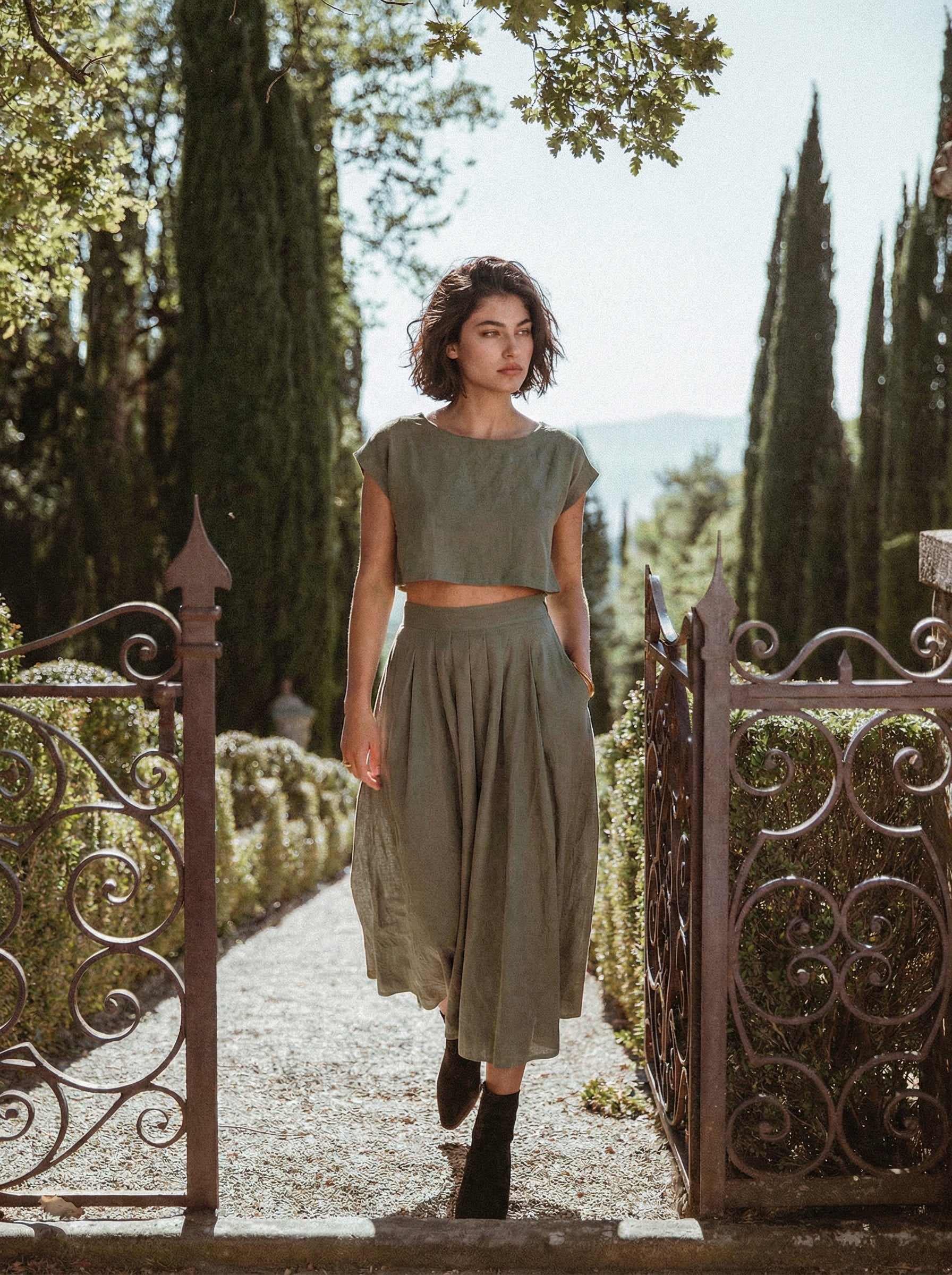 Model wearing the Sonfre NAOMI eucalyptus green linen two-piece set, featuring a boxy crop top and a high-waisted midi skirt, walking through an ornate iron gate in a garden setting.