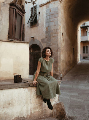Model wearing the Sonfre NAOMI eucalyptus green linen crop top and high-waisted midi skirt set, leaning against an ornate stone balcony railing with trees in the background.