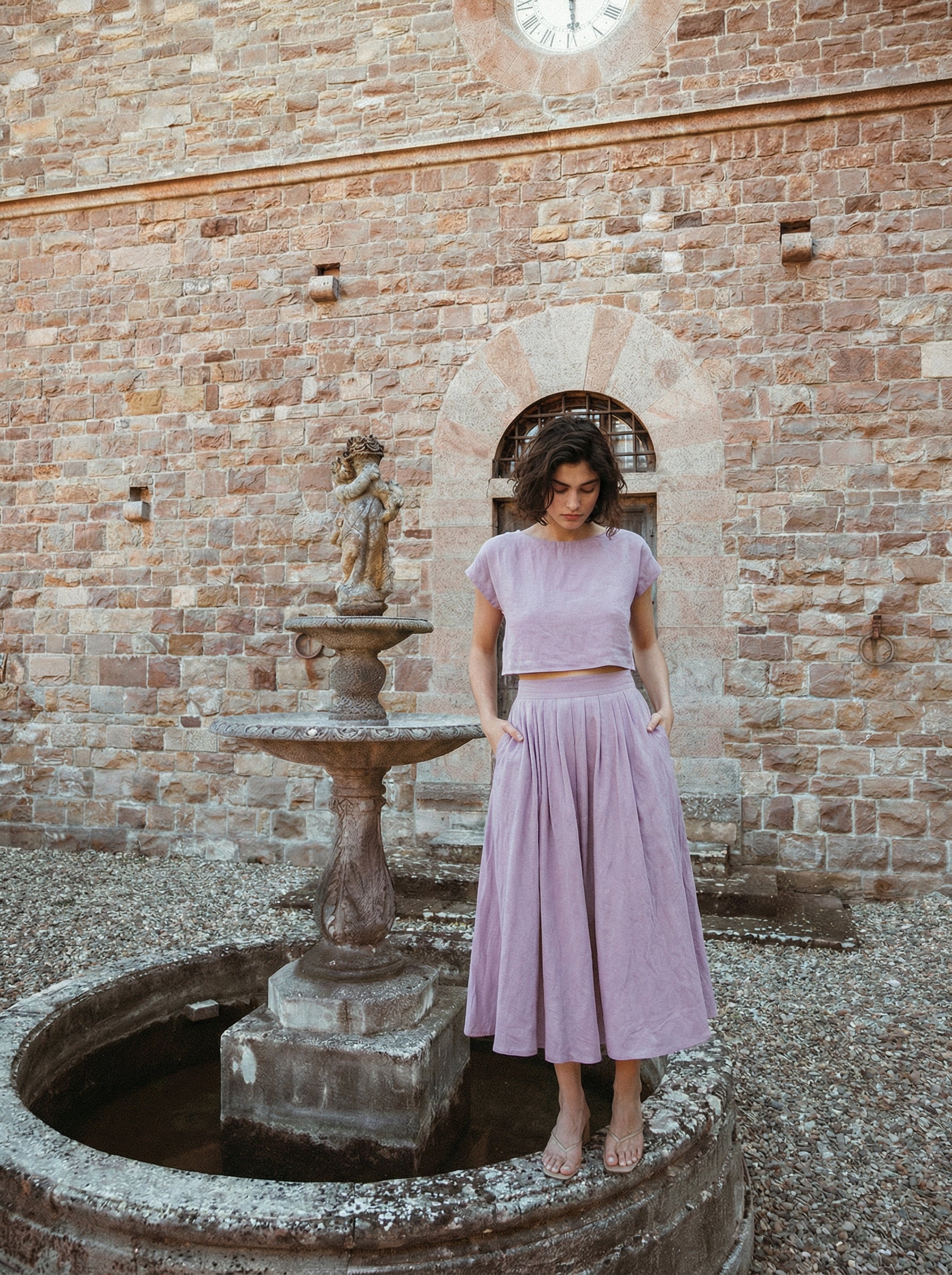 Model wearing the Sonfre NAOMI lilac linen two-piece set, featuring a cropped top and a high-waisted midi skirt, standing next to an old stone fountain against a brick wall.