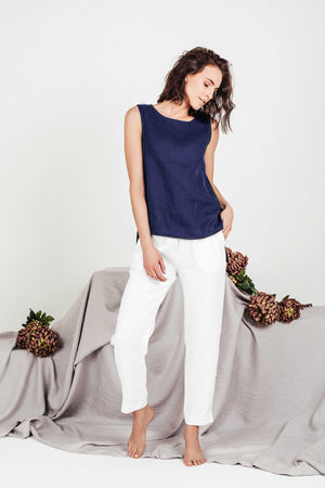 Woman in navy linen top and white linen pants, gently looking down with relaxed posture, barefoot on a soft fabric backdrop decorated with large dried flower bunches.