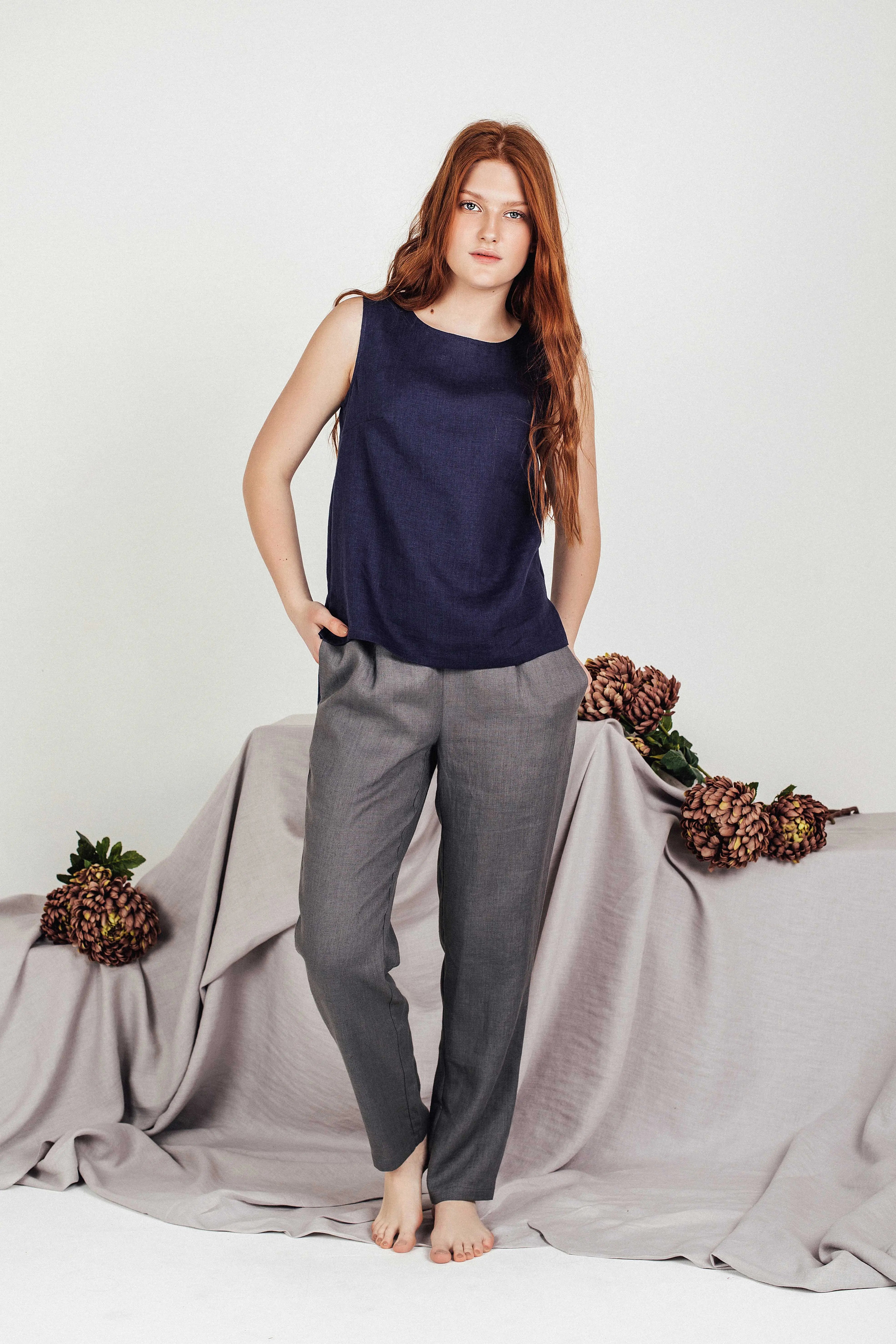 Red-haired woman in a navy blue linen sleeveless top and high-waisted grey pants, barefoot with a soft expression, styled against a neutral draped fabric backdrop with dried flowers.