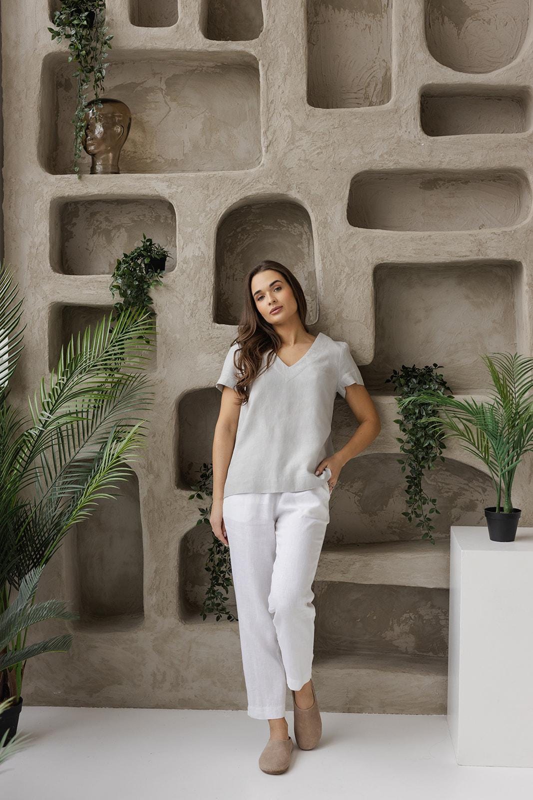 Model in white linen pants and a gray top, leaning against a neutral-toned wall with green plant decor, emphasizing the natural, lightweight, and breathable fabric