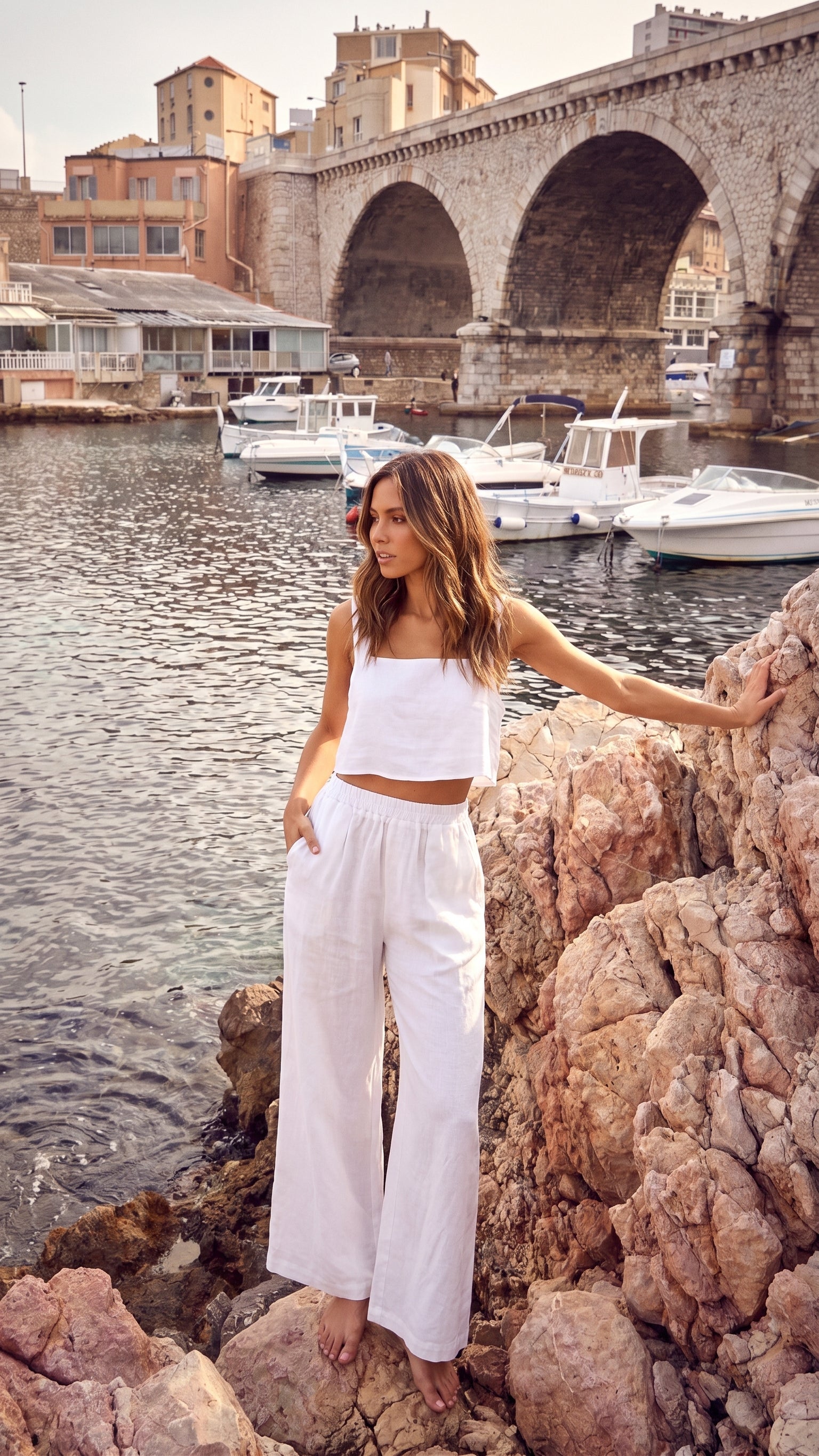 Model standing on rocky terrain wearing the TORA white linen cropped top, with a boat harbor and arched stone bridge in the background.