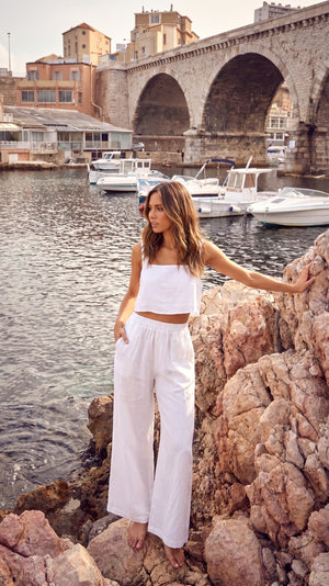 Model standing on rocky terrain wearing the TORA white linen cropped top, with a boat harbor and arched stone bridge in the background.