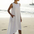 Woman wearing a sleeveless white linen dress with an asymmetrical high-low hem and side pockets on the beach.