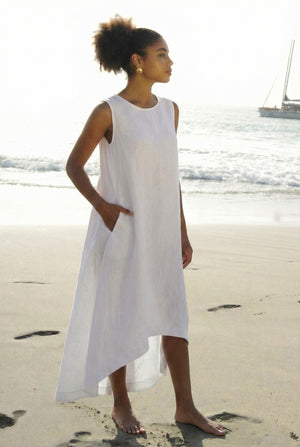 Woman wearing a sleeveless white linen dress with an asymmetrical high-low hem and side pockets on the beach.