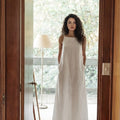 Woman standing in doorway wearing minimalist white linen maxi dress with square neck and pockets.