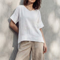 Model wearing an oversized white linen tunic top with short sleeves, leaning against a sunlit concrete wall.