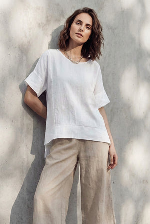 Model wearing an oversized white linen tunic top with short sleeves, leaning against a sunlit concrete wall.