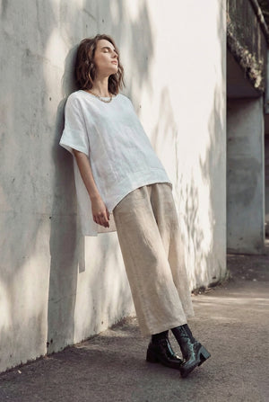 Woman leaning against a concrete wall wearing an oversized white linen tunic, wide-leg beige linen pants, and black leather boots.