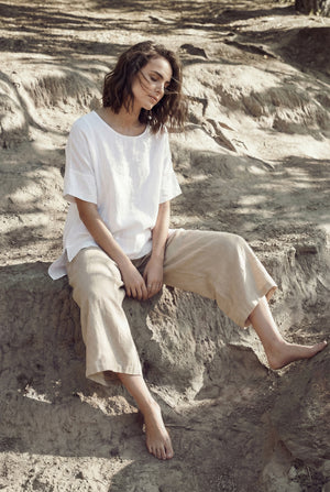 Woman sitting outdoors on a sandy bank wearing a relaxed white short-sleeve linen tunic and beige wide-leg linen trousers.